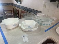 Overview of the 11 pieces grouped on kitchen counter: Pyrex white dishes with blue Cornflower pattern, and clear glass bowls and casserole dish with lid.