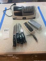 Photo of Smiths electric cutlery sharpener, 4 knives with black handles, two pocket tools, all items on a countertop.