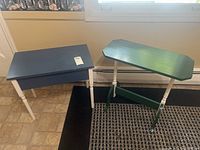 Two wooden side tables: one blue rectangular with white legs and drawer, one green asymmetrical with white legs and green crossbar.
