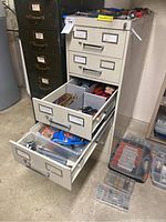 Beige multi-drawer storage cabinet filled with hand tools and some plastic bags of wire connectors, shot showing multiple open drawers with tools inside, along with plastic organizer boxes at the base.