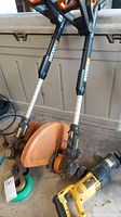 Two Worx WG160 trimmers standing upright with orange guard covers, visible adjustable shafts, positioned next to a Dewalt reciprocating saw laying on the ground. Trimmers missing batteries and appear used.