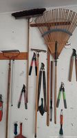 Wall-mounted yard and garden tools including various rakes, brooms, pruners, and squeegee arranged on wooden racks