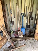 Collection of multiple yard and garden tools stored against a wooden wall including shovels, pickaxes, an edger, and a broom.