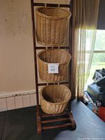 Full view of three woven wicker baskets vertically hanging from dark brown wooden ladder-style stand on wheels beside window.