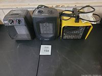 Photo of three electric fan heaters arranged side by side on a table: gray heater, black 'Classic' heater, and yellow heater with handle.