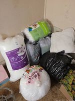 Photo showing five large bags of white batting and four white square pillow forms stacked in a corner.