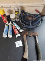 Photo showing booster cables, various tubes and spray cans of sealants, two hand tools, and two metal hatches on a black surface against a white wall.