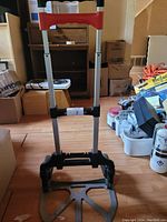 Full view of the collapsible dolly showing the upright handle with red grip and the base platform on the floor.