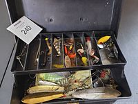 Open tackle box showing upper tray with assorted fishing lures in compartments and loose lures beneath