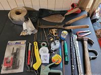 Overview of assorted hand tools and hardware items on black surface, showing a masonry trowel, two hammers, piano hinges, staple gun, tape measures, and small parts.