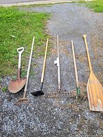Photo showing six gardening tools laid on a gravel surface: round mouth shovel, hoe, lawn rake, garden rake, hand rake, and wooden paddle.