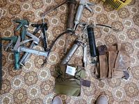 Five caulking guns, four grease guns, and two leather tool belts displayed on patterned tile floor. The tools show signs of wear and use.