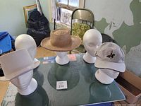 Image shows five white mannequin heads on a glass table, three with hats (white top hat, straw hat, baseball cap). Background includes chair and peeling plaster wall.