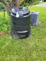 Large green compost bin placed outdoors on grass next to a tree and another plastic bin