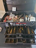 Open black plastic tackle box showing multiple compartments filled with fishing tackle including bobbers, metal lures, hooks, and lines.