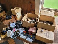 Wide view showing several boxes and bags containing assorted hardware including hinges, nails, screws, and other items on a wooden table.