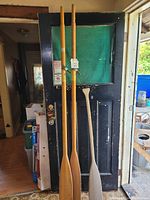 Photo showing two wooden oars and one wooden paddle hung on a black door in an indoor area with some surrounding clutter.