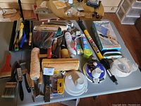Overview of the lot showing paint rollers, brushes, paint can lids, scrapers, sanding tools and more spread on a table.
