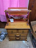 Photo showing a very large roll of plastic wrap mounted on a wooden spindle stand placed on top of a small oak nightstand with multiple drawers.