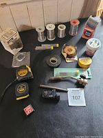 Photo showing five rolls of solder wire, tubes of solder, Kester solder box, soldering paste container, and some tape measures on a black surface.