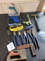 Photo showing a set of hand tools including 5 blue-handled pliers, a hammer with a rubber grip, an orange and silver staple gun, a yellow ratcheting strap, assorted drill bits, a plastic blow gun with nozzle, and a circular saw blade on a black surface.