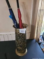 Photo of a brass umbrella stand with embossed floral decoration, holding a blue umbrella and a red umbrella.