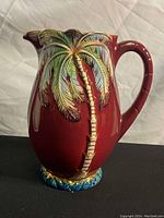 Front view of the red ceramic pitcher featuring a raised palm tree design with green and gold accents and textured blue and gold base.