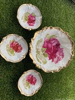 All four bowls laid out together on grass showing large bowl and the three smaller bowls, displaying the hand-painted pink rose motifs and gold scalloped edges.
