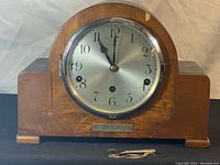 Front view of wooden Art Deco mantle clock with walnut veneer and silver clock face with Arabic numerals. Small winding key placed in front of clock.