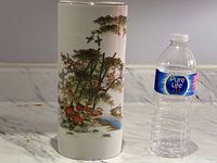 Full view of the tall cylindrical porcelain vase next to a water bottle to show scale. The vase features a hand painted nature scene with birds, bamboo, flowers, waterfall on a white base.