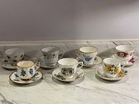 Full view of the seven porcelain teacups and saucers arranged in a row, showing the variety of floral designs and gold trim.