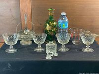 Photo showing the full lot: four clear glass dessert cups with pedestal bases, a vintage glass serving basket with metal handle and spoon, green glass decanter with gold floral decoration, a clear glass measuring cup, and a small cut glass bottle with stopper, alongside a water bottle for sizing.