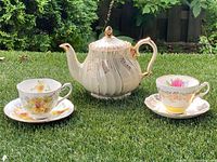 Lot of a white ceramic teapot with vertical gold stripes and floral detailing, gold accented handle and lid, set on grass with two teacups and matching saucers with floral patterns in yellow and pink.
