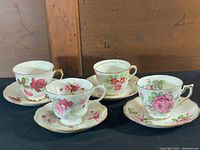 Four vintage floral teacups with matching saucers, decorated with roses and flowers, gold trim visible, arranged in a row on a wooden surface.