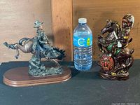 Native porcelain decanter shaped as a Native American horse and rider and a bronze-colored cowboy cowboy figurine on a wooden base with a water bottle for scale.
