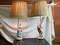 Pair of vintage porcelain figurine table lamps with brass bases and pleated fabric shades, shown lit and side by side on a table.