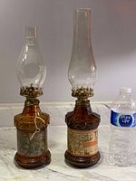 Full view of both vintage oil lamps with water bottle for scale, showing amber glass bases with decorative map labels and clear glass chimneys with brass fittings.
