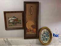 Photo showing three picture frames: a square wooden frame with needlepoint house art, a tall rectangular frame with still life painting, and an oval frame with floral painting beside a water bottle for scale.