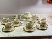 Full arrangement of tea cups and saucers with teapot, sugar bowl, creamer, and handled bowl displayed on marble counter.