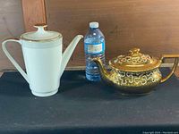 Front view of the white ceramic coffee pot and gold-tone teapot next to a water bottle for scale.