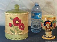 Photo showing vintage ceramic cookie jar with floral design and green lid beside vintage ceramic wine mug with sculpted figure and grapevine decoration, alongside a water bottle for size reference.