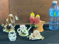 Photo showing six vintage ceramic decorative figurines including birds on branches, a bear-shaped teapot, and floral baskets with a water bottle for scale.