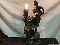 Full view of the vintage table lamp with figurines of fisherman and woman, light bulb illuminated showing decorative leaf accents.