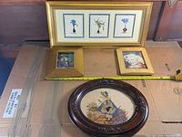 Group photo showing four vintage picture frames on a flat surface, including large gold frame with three botanical prints, two smaller gold-tone frames with photos, and one oval dark wood frame with stitched artwork.
