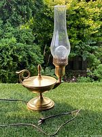Full view of the vintage solid brass table lamp on grass background showing lamp base, lid, finial, electrical cord, and glass chimney.