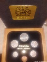 Wooden display box containing six Canadian coins arranged in black velvet interior. Gold Canadian coat of arms plaque mounted inside lid.
