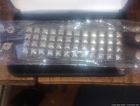 Black leather bracelet band with rows of silver pyramid studs inside clear plastic packaging, shown laid flat in a box.