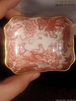 Porcelain dish held in hand to demonstrate size and gold-edge detail with red bird and flower design.