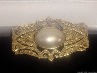 Close-up of gold-tone brooch with oval faux pearl centerpiece showing intricate filigree detailing.