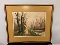 Framed etching print surrounded by a golden-beige mat and wooden frame showing a canal lined with trees in autumn, with walking figures.